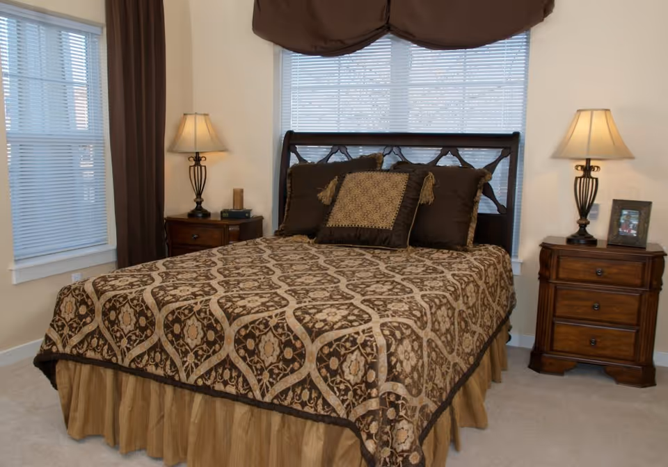 A neatly made bed with patterned bedspread, decorative pillows, two bedside tables and lamps in a bright bedroom with windows.