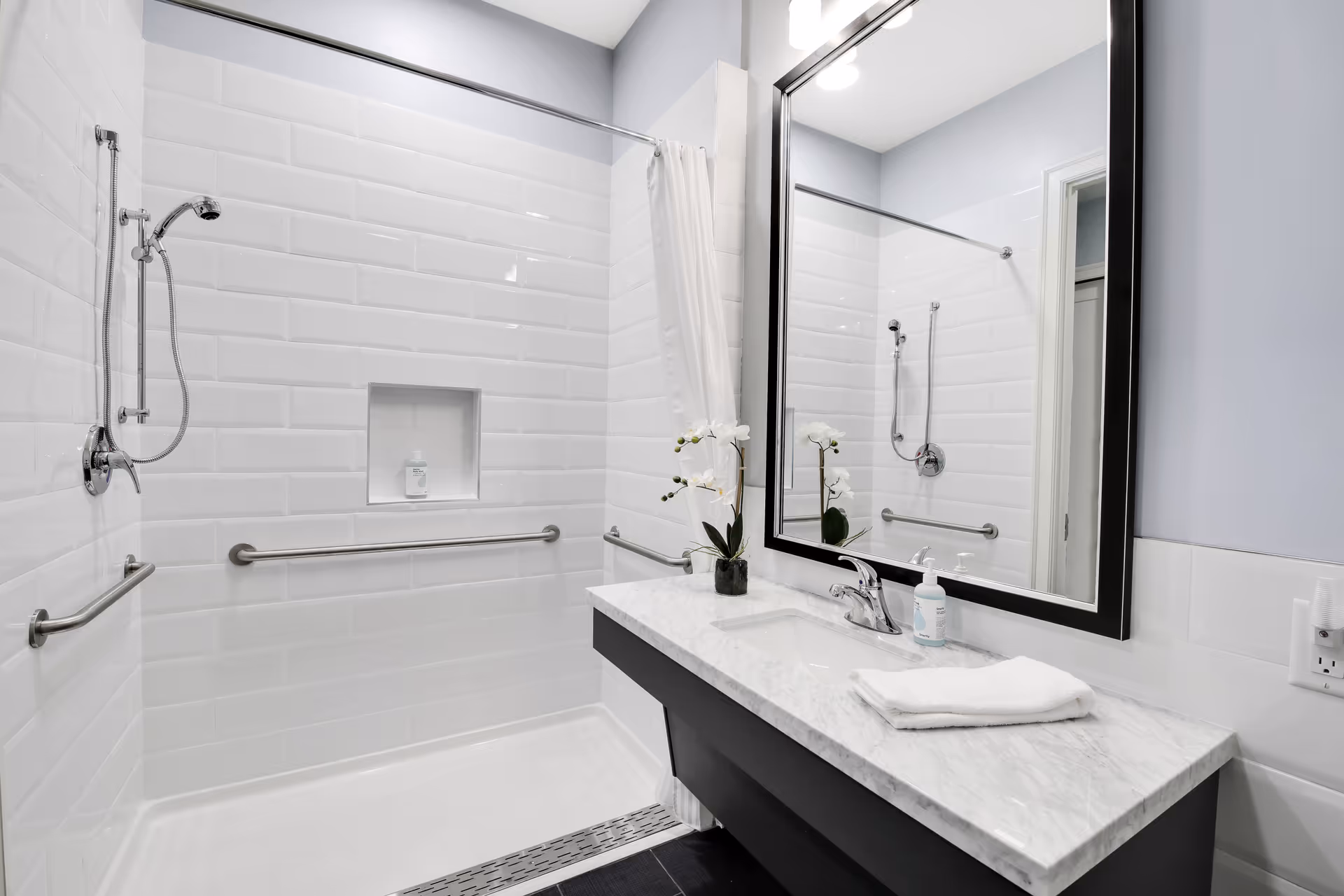 A modern bathroom with white subway tile walls, a walk-in shower with a handheld showerhead, grab bars, and a built-in niche holding a bottle. There is a white shower curtain on a rod. A vanity with a marble countertop, a sink, a faucet, a small potted orchid, a bottle of hand soap, and a folded white towel is visible. A large rectangular mirror with a black frame hangs above the vanity.