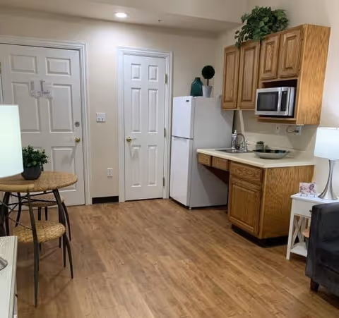 Interior view of a small kitchen area with wooden cabinets, a white refrigerator, a microwave, a sink, and a countertop. There are two closed white doors on the left side of the image. A small round table with two chairs and a potted plant is visible on the left, and a white side table with a lamp and a small photo frame is on the right. The floor is wooden, and the walls are light-colored.