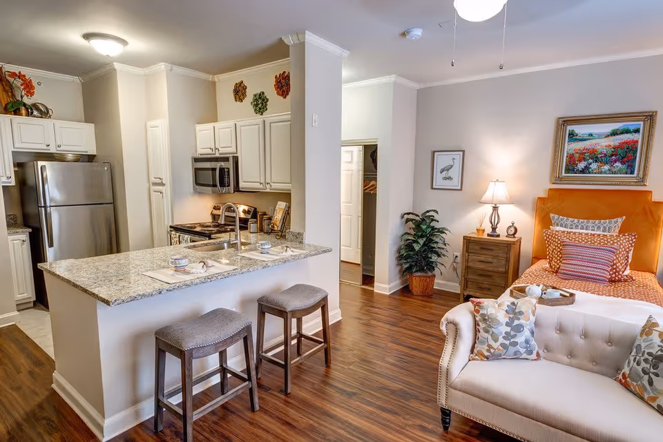 A cozy studio apartment interior featuring a small kitchen with white cabinets, stainless steel refrigerator, microwave, and granite countertop with two stools. Adjacent to the kitchen is a living and sleeping area with a beige tufted sofa, a bed with an orange headboard and patterned pillows, a wooden nightstand with a lamp, and framed artwork on the walls. The floor is wood, and there is a potted plant near the nightstand.