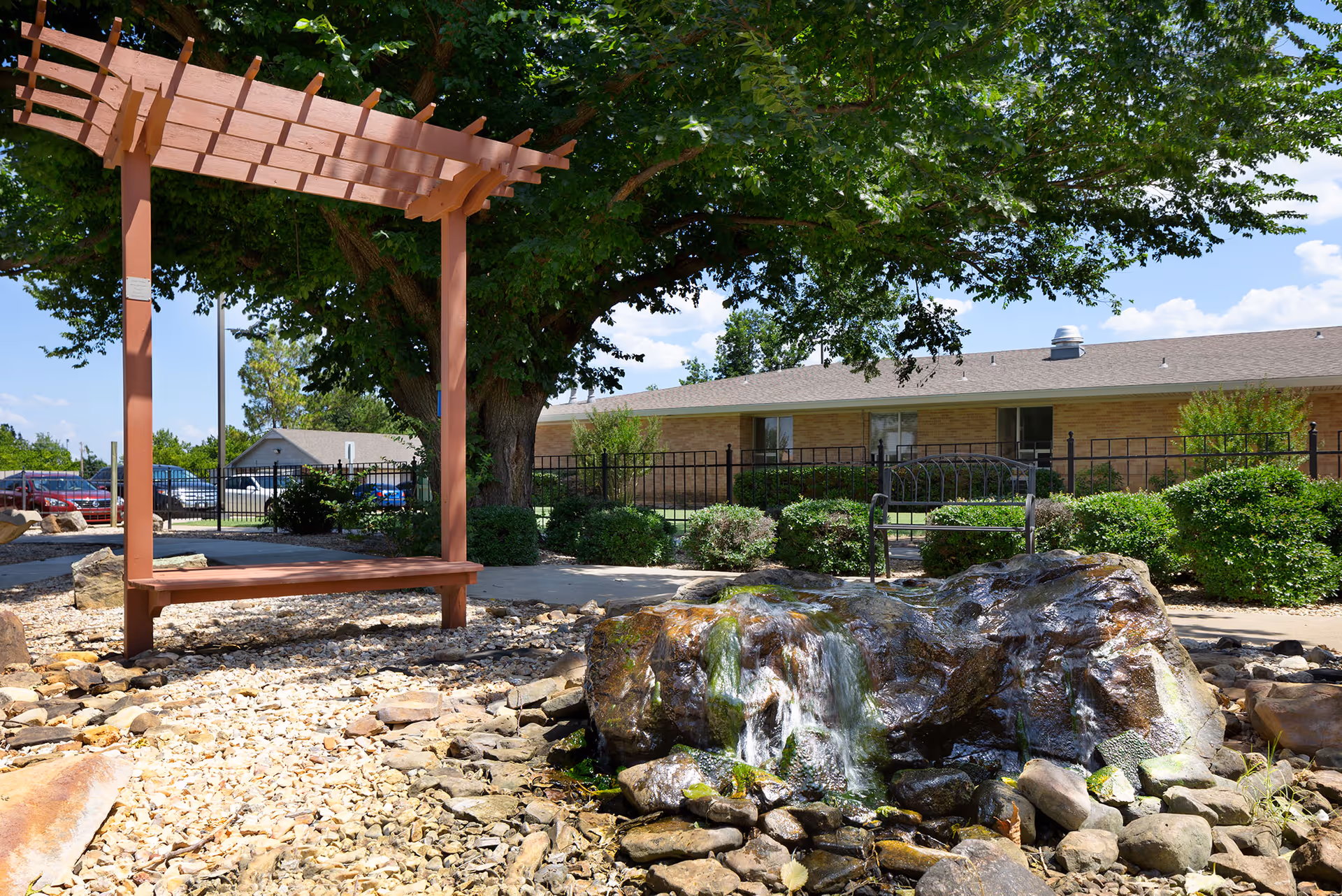 Sunny outdoor courtyard with a wooden pergola bench, a small rock waterfall feature, and a single-story brick building in the background.