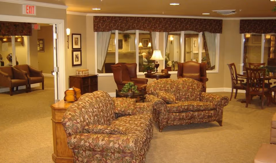 A cozy living room area in a senior care facility featuring two floral-patterned sofas, two brown leather armchairs, a wooden side table with a lamp, and a small round table with chairs near the window. The room has beige walls and carpet, with framed pictures on the walls and an exit sign above a doorway.