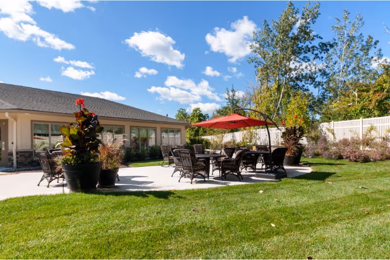 Outdoor patio area at Viva Memory Care at Dresher with several tables and chairs arranged on a concrete surface. One table has a large red umbrella providing shade. There are large potted plants around the patio and a white fence enclosing the area. The sky is blue with scattered clouds and trees are visible in the background.