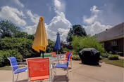 Outdoor patio area with a round glass table surrounded by colorful chairs in red, blue, and purple. Two large umbrellas, one yellow and one blue, provide shade. A black covered grill is visible on the concrete patio. Green bushes and trees surround the area under a partly cloudy sky.