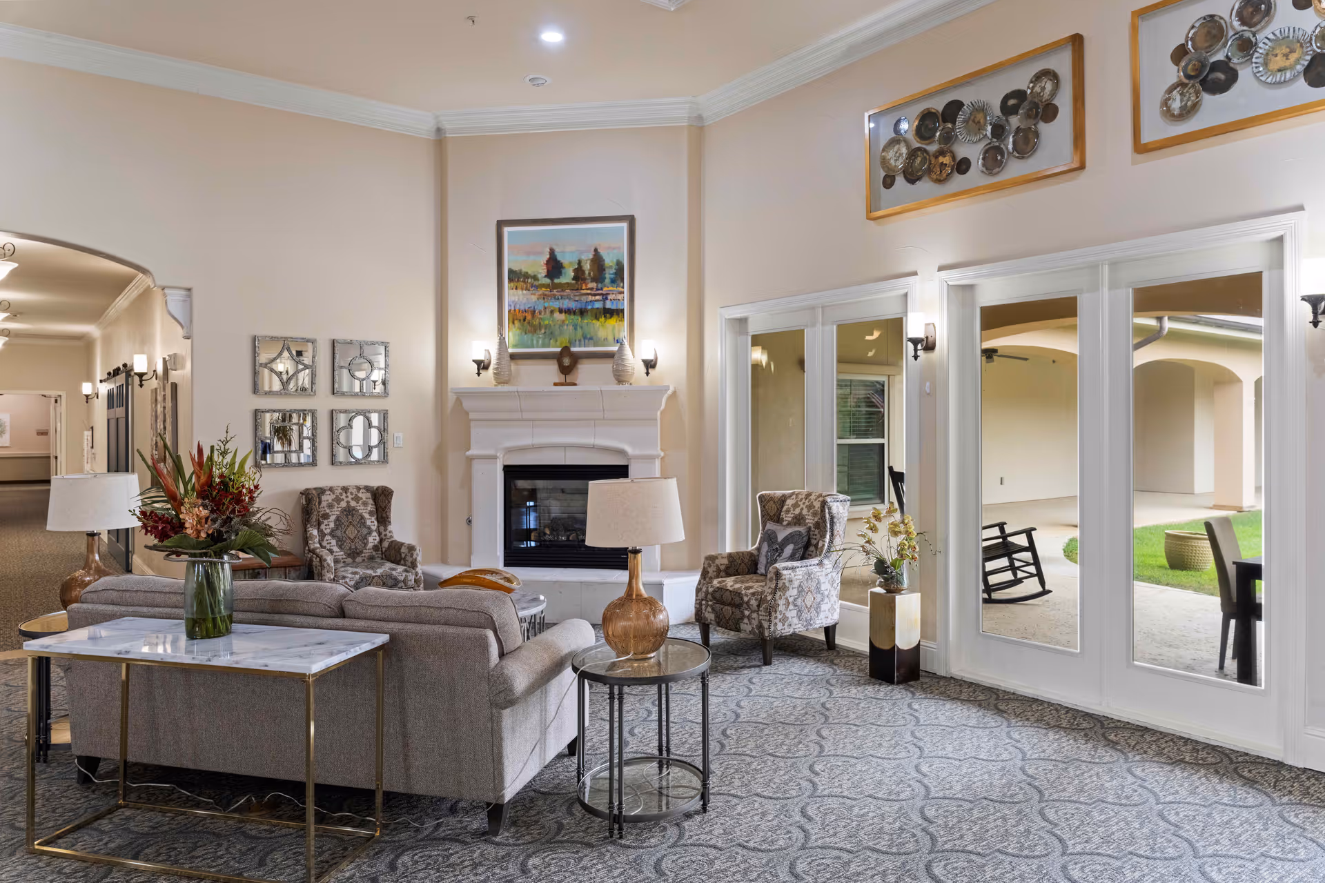 A cozy senior living common area with a beige sofa, two patterned armchairs, a fireplace with a painting above it, and large glass doors leading to an outdoor patio with rocking chairs and a table. The room is decorated with lamps, a flower arrangement, and wall art, featuring a patterned carpet and soft lighting.