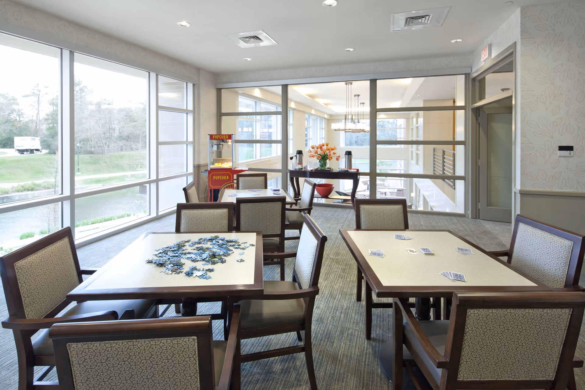 A bright common area with large windows overlooking greenery and a waterway. The room contains several tables and chairs, with one table having a partially completed jigsaw puzzle and another table with playing cards spread out. In the background, there is a popcorn machine and a table with coffee dispensers and a vase of flowers.