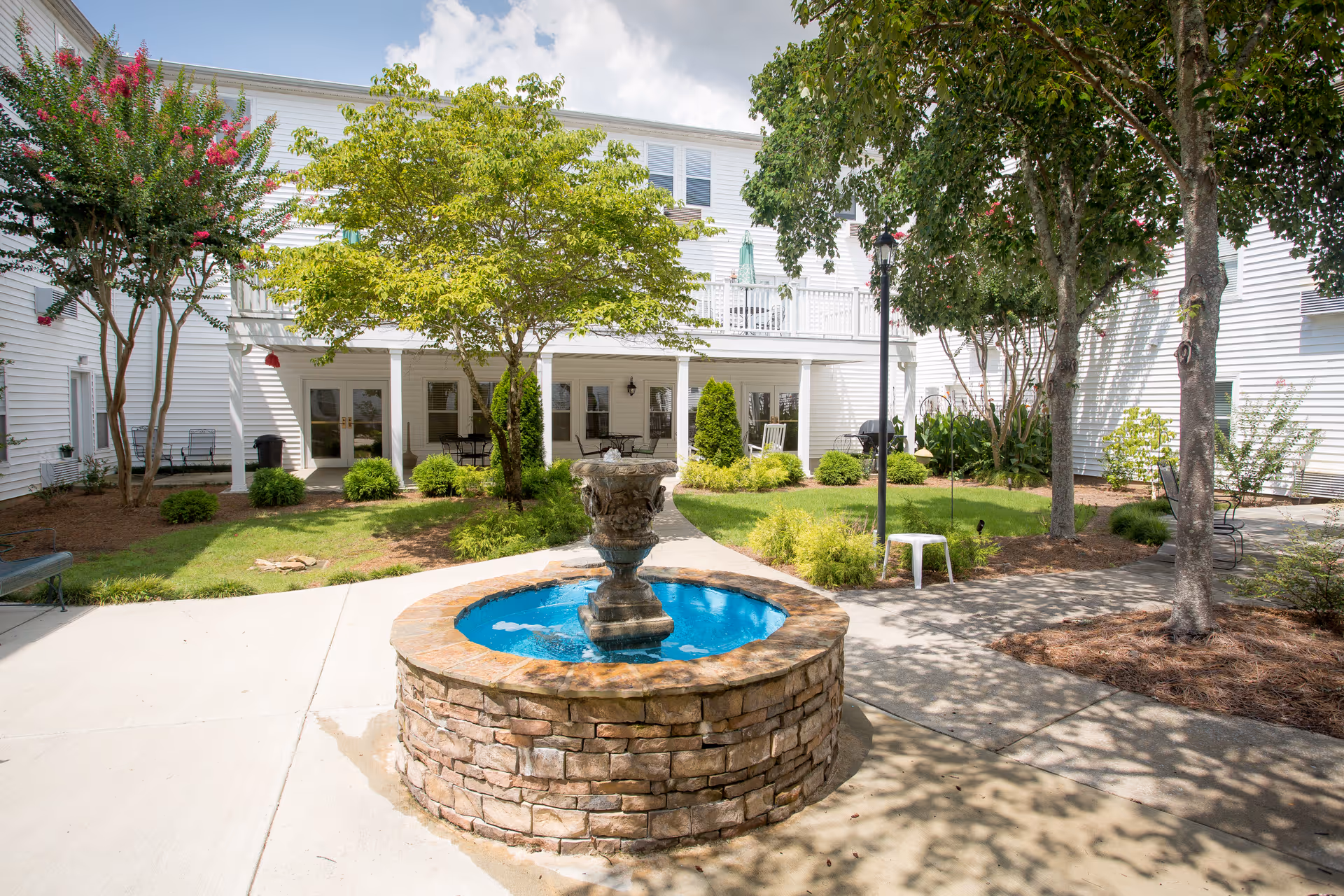 Outdoor courtyard area at Morningside of Vestavia Hills featuring a stone fountain with blue water in the center, surrounded by trees, shrubs, and a paved walkway. The white building with multiple windows and a covered patio with seating is visible in the background under a partly cloudy sky.