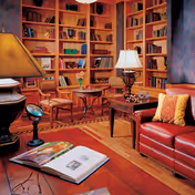 Cozy living room with wooden bookshelves filled with books, a red leather sofa with a yellow pillow, a wooden coffee table with a lamp, and two wooden chairs around a small round table. There is also an open book on a wooden table in the foreground and a floor lamp on the left side.