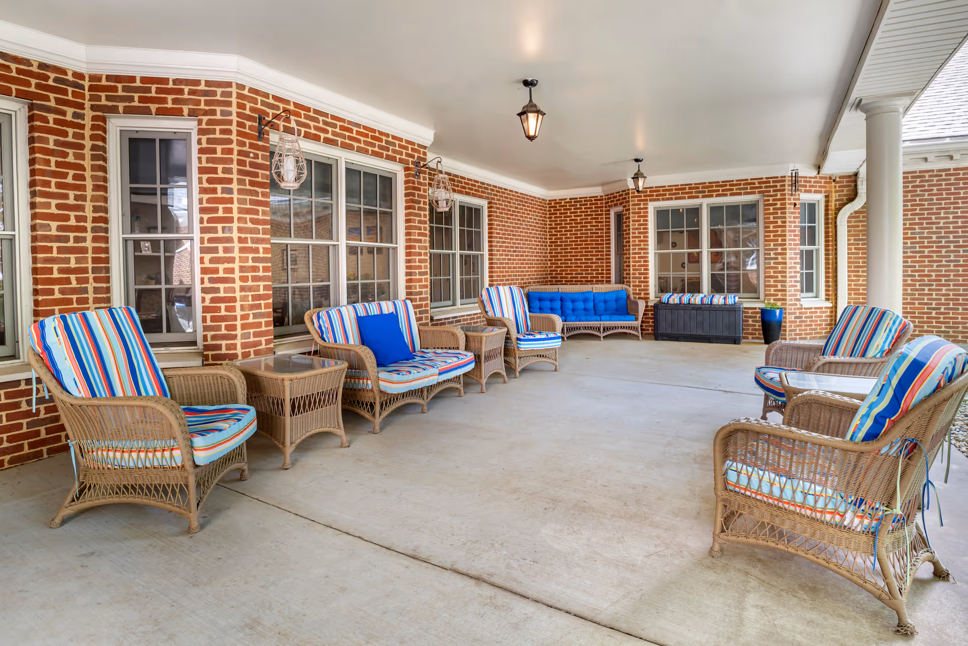 Covered outdoor patio area with brick walls and multiple wicker chairs and sofas with colorful striped cushions and blue pillows. The space has hanging lantern-style lights and large windows along the walls.