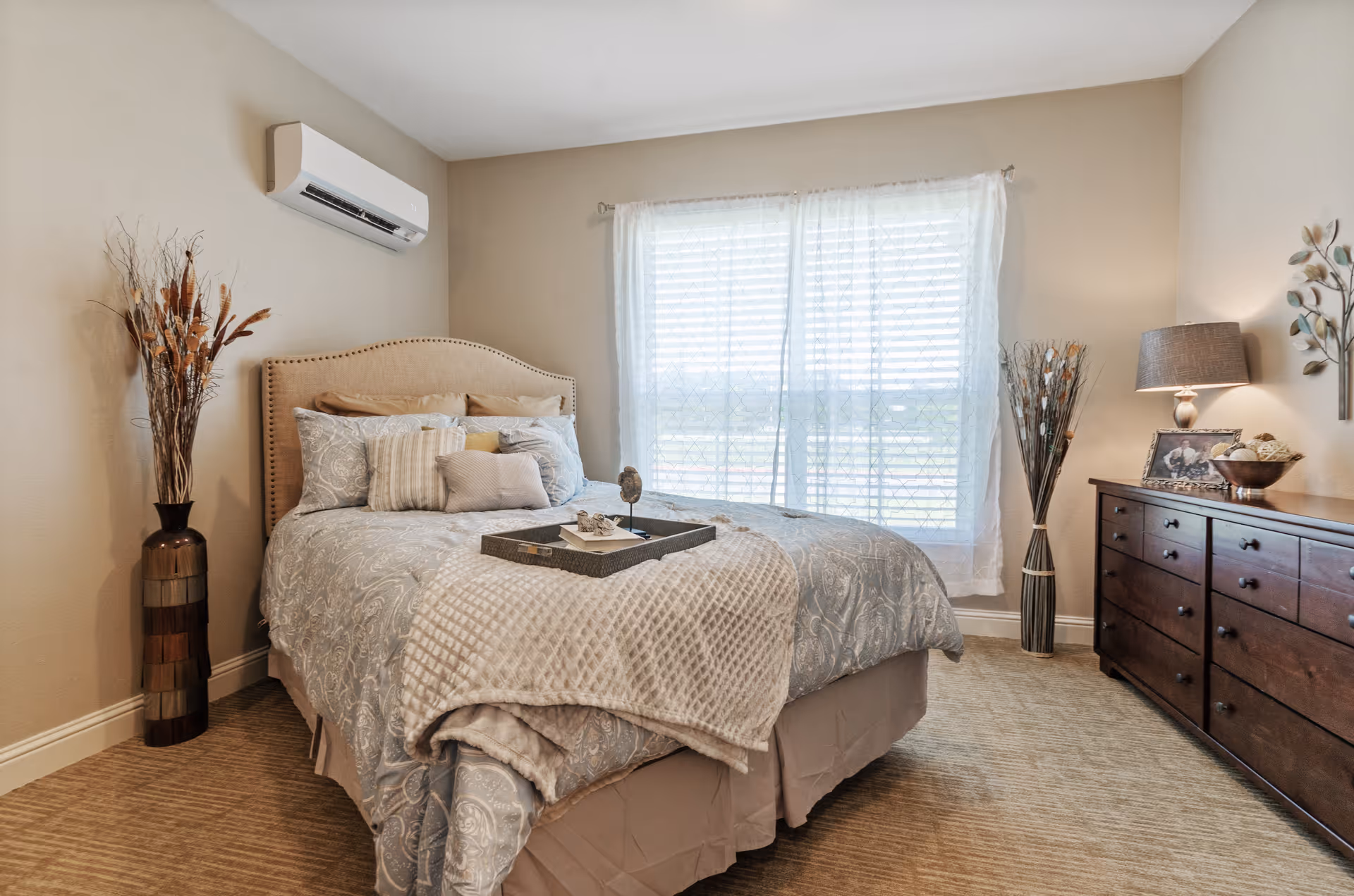 A neatly made bedroom with a upholstered bed, decorative pillows and throw, a wooden dresser with a lamp, vases, and a window with sheer curtains.