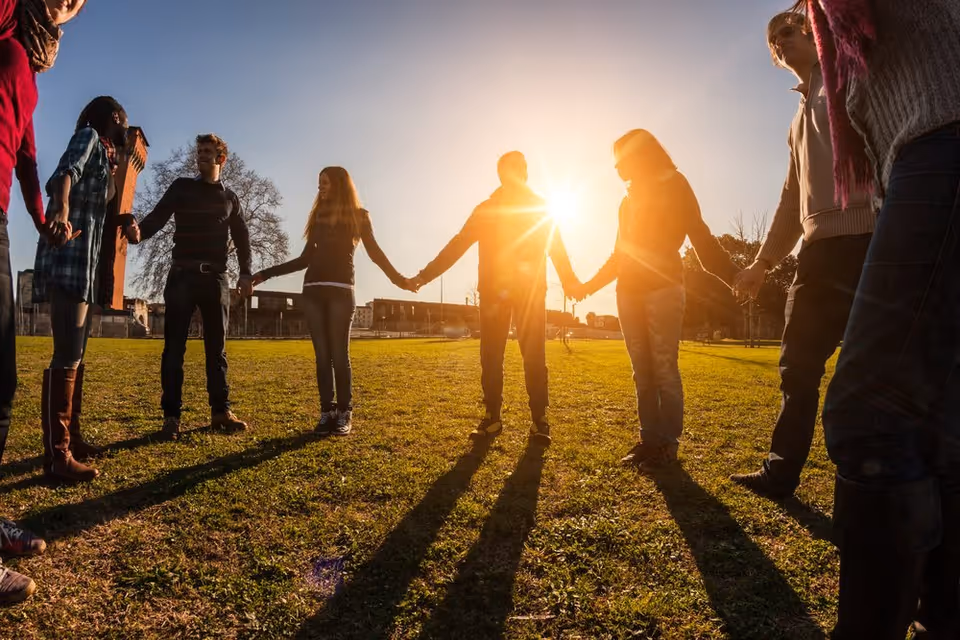 A group of people standing in a circle holding hands outdoors on a grassy field with the sun shining brightly in the background, casting long shadows.