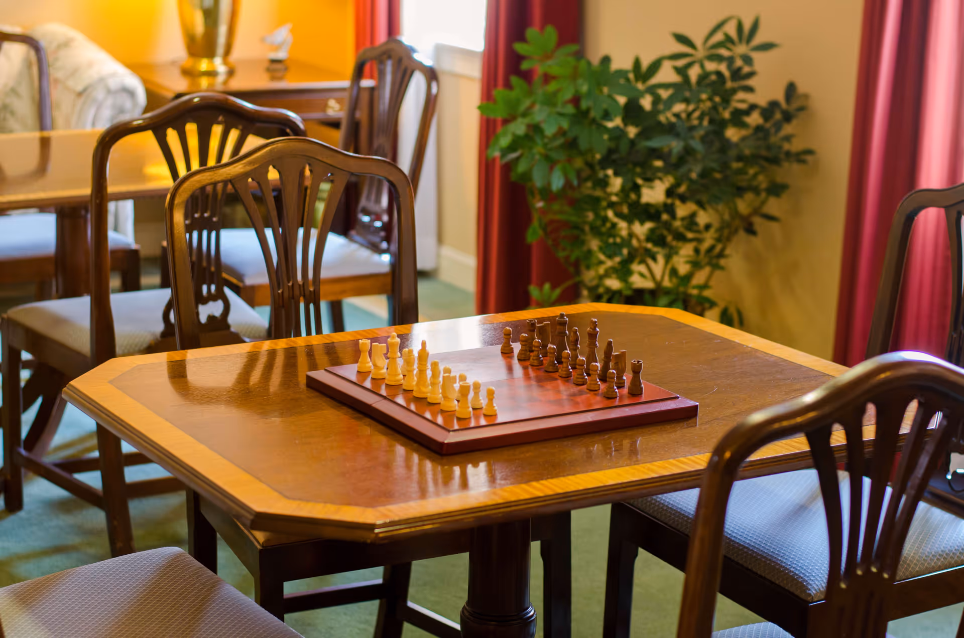 A wooden table with a chessboard set up for a game, surrounded by wooden chairs with cushioned seats. In the background, there is another table with chairs, a potted plant, and red curtains near a window.