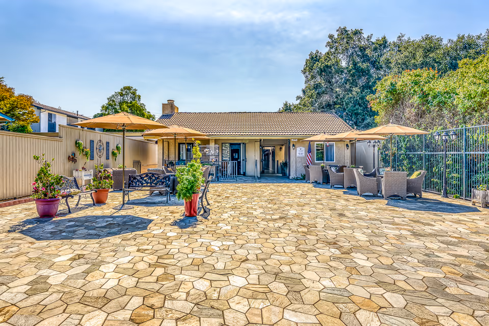 Outdoor patio area with stone tile flooring, several seating arrangements including benches and wicker chairs with umbrellas, potted plants, and a single-story building with a tiled roof in the background under a clear blue sky.