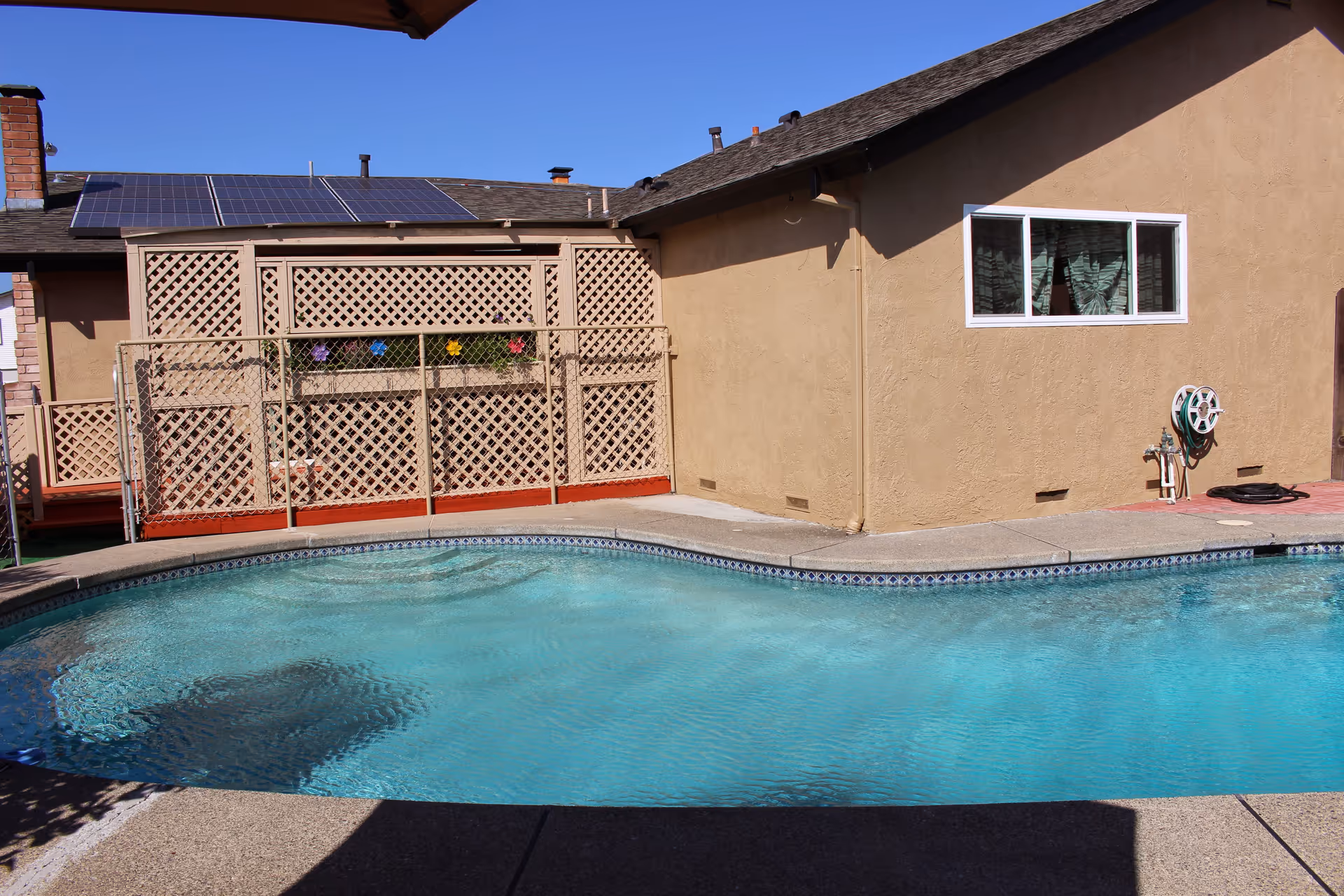 Outdoor swimming pool next to a beige stucco building with a window and a garden hose reel mounted on the wall. There is a wooden lattice fence with colorful flower decorations and solar panels on the roof under a clear blue sky.