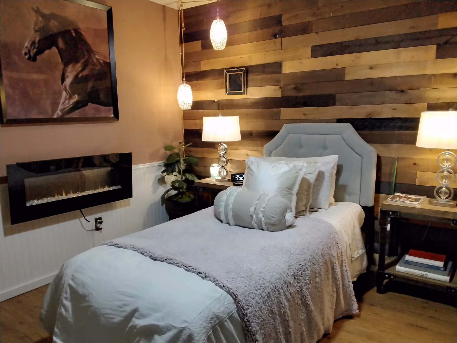 A cozy bedroom with a single bed featuring a light gray upholstered headboard, white bedding, and multiple pillows. The room has a wooden accent wall behind the bed, two bedside tables with lamps, a digital alarm clock, a potted plant, a wall-mounted electric fireplace, and a framed picture of a horse on the adjacent wall.