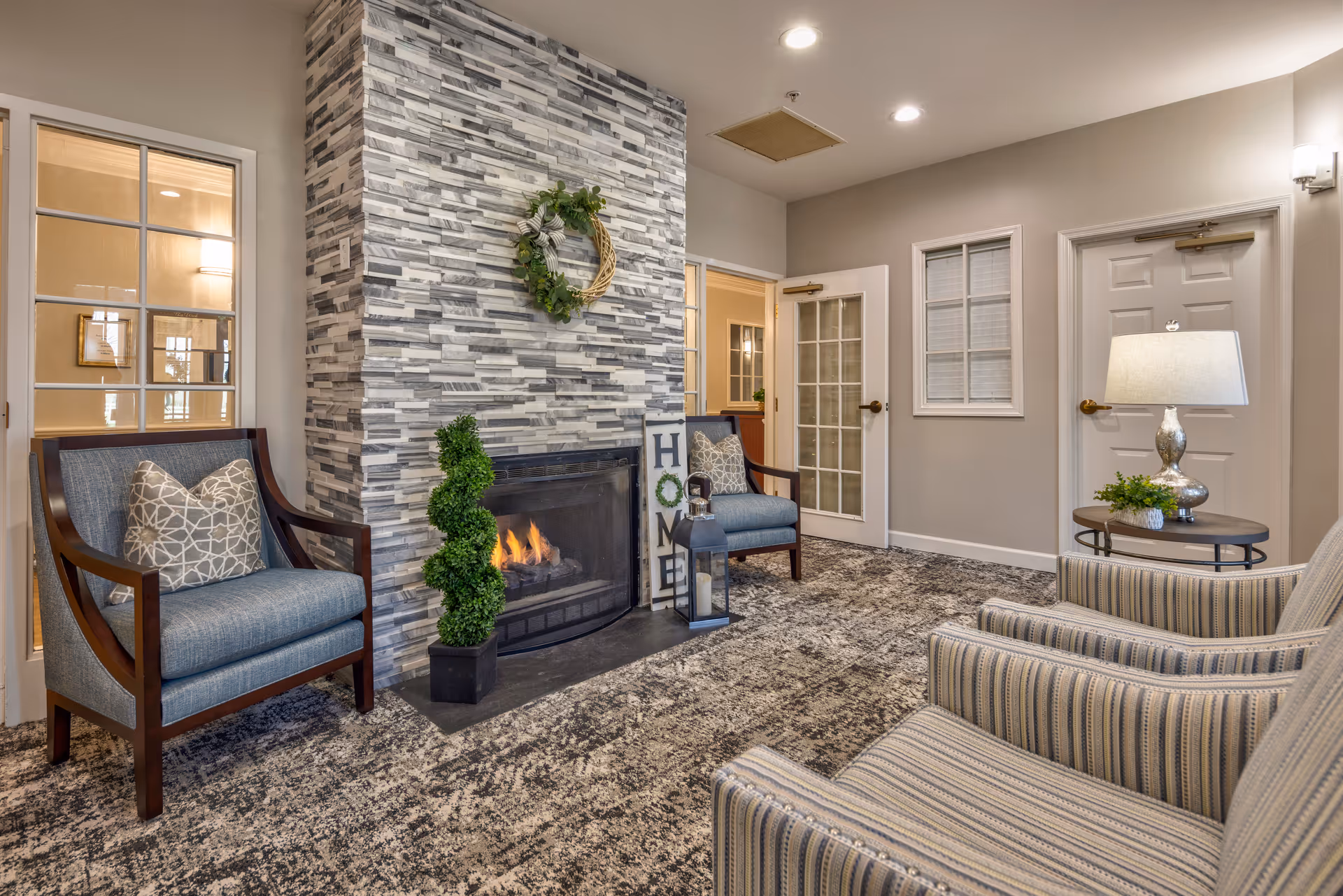 A cozy living room area with a modern stone fireplace in the center, decorated with a green wreath and a 'HOME' sign. Two blue cushioned wooden armchairs are placed on either side of the fireplace, and two striped armchairs face the fireplace. A small round side table with a lamp and a plant is next to the striped chairs. The room has neutral-colored walls, carpeted floor, and multiple doors and windows.