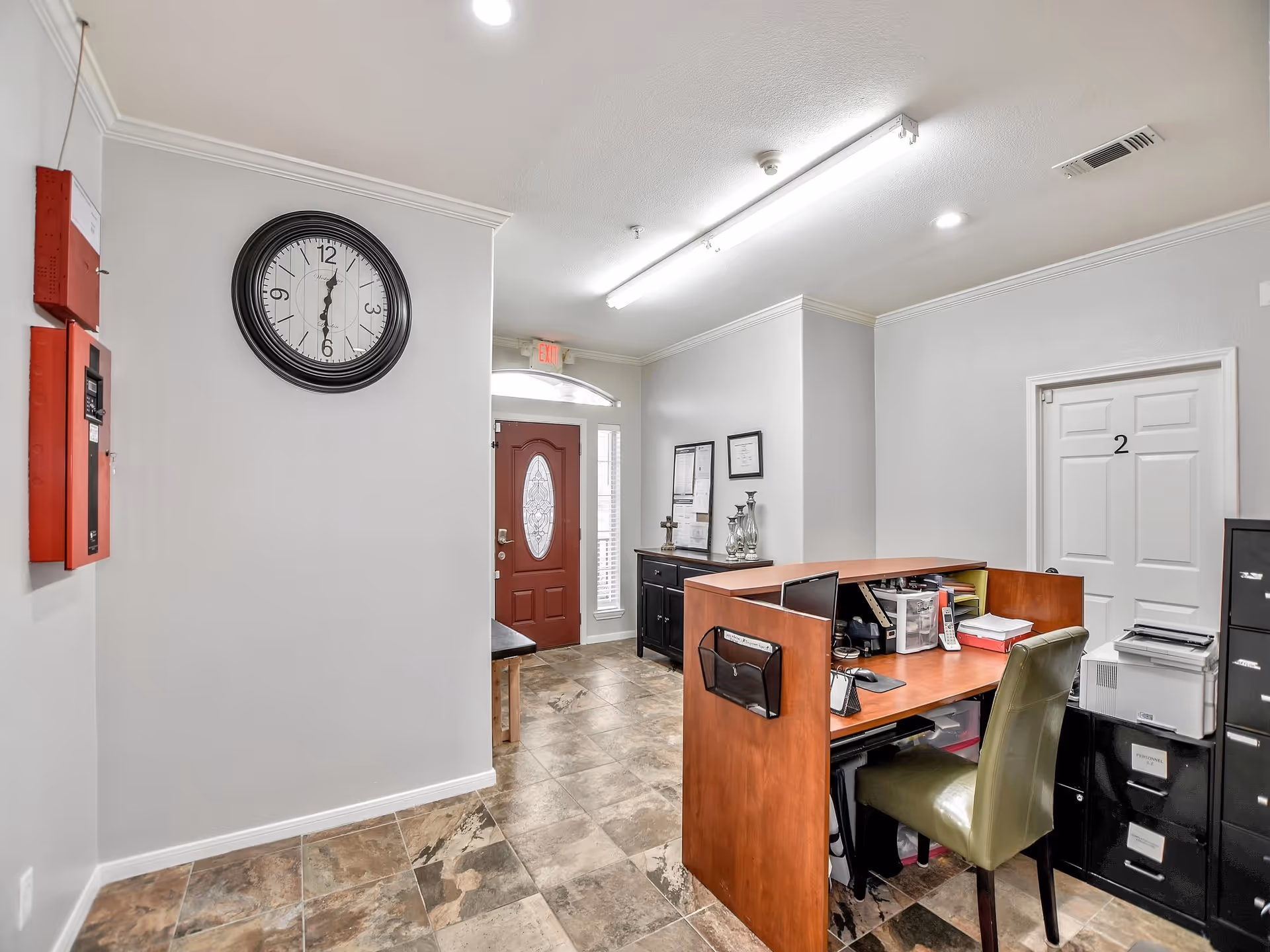 Interior view of a reception or office area in an assisted living facility. The space features a wooden desk with office supplies, a green chair, a black filing cabinet, and a door labeled with the number 2. There is a large round wall clock showing the time as 6:30, a red front door with decorative glass, and tiled flooring.