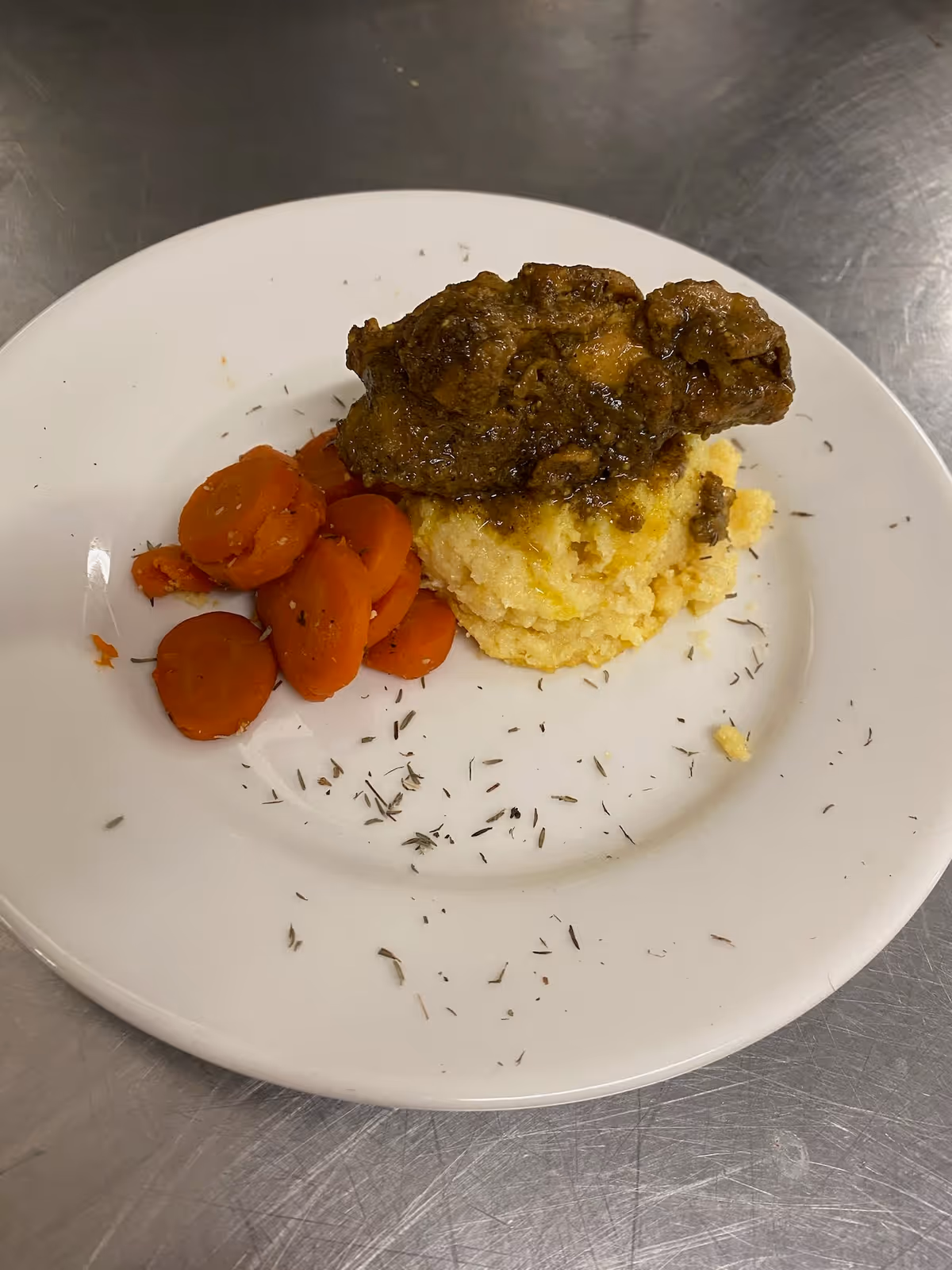 A white plate with cooked sliced carrots on the left side and a serving of mashed potatoes topped with a piece of seasoned meat on the right side, garnished with herbs scattered on the plate. The plate is on a metallic surface.
