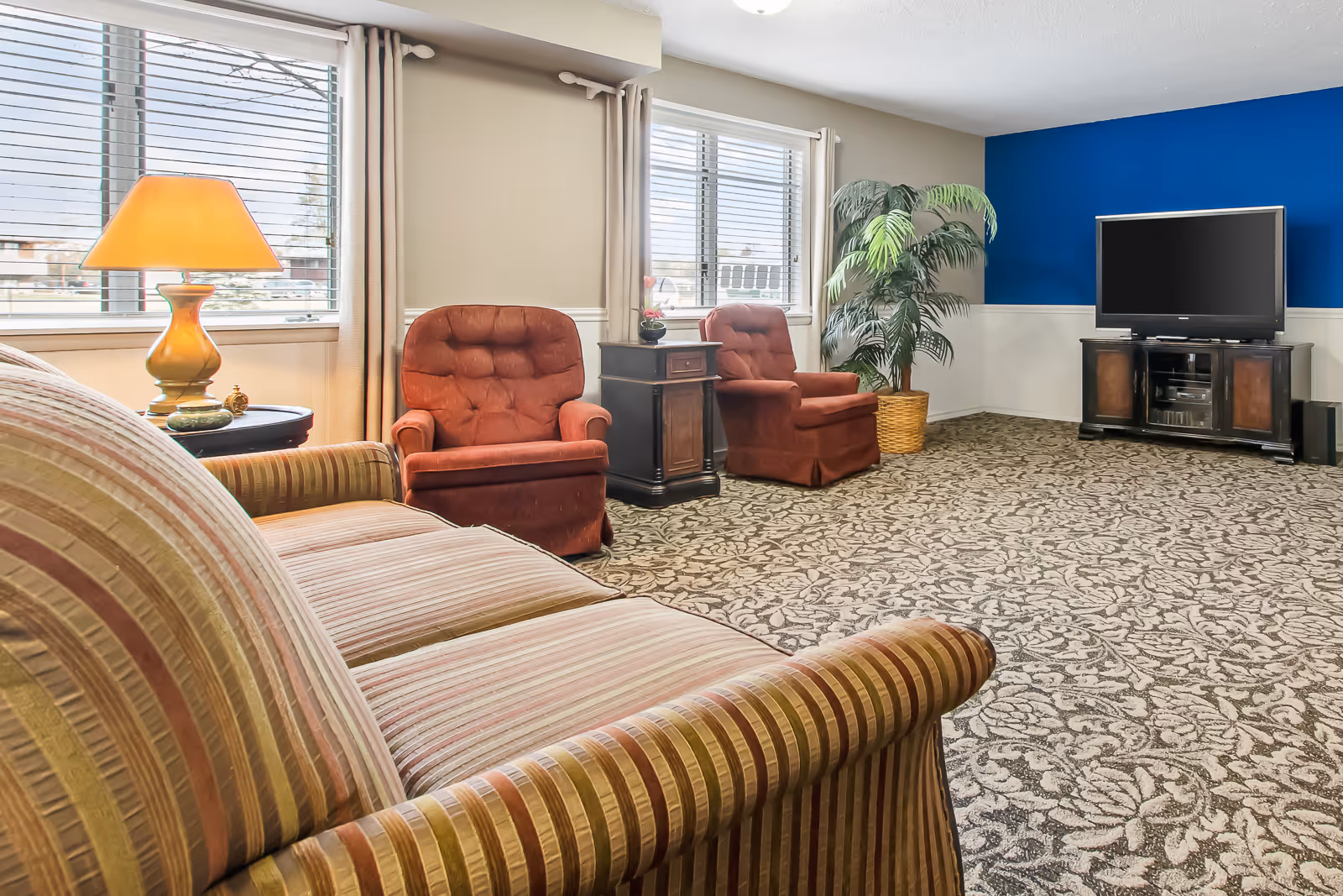 Communal living room with a striped sofa in the foreground, two red armchairs by windows, a TV on a stand, and a lamp.