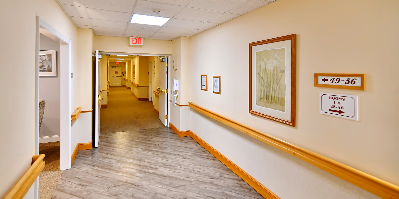 Interior hallway of a senior living facility with light-colored walls, wooden handrails, framed artwork, directional signs for room numbers, and an exit sign above double doors leading to another corridor.