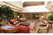 A spacious and well-lit living room area with comfortable seating including sofas and armchairs arranged around a coffee table on a patterned rug. There are large indoor plants, table lamps, and a high ceiling with skylights allowing natural light to fill the space.