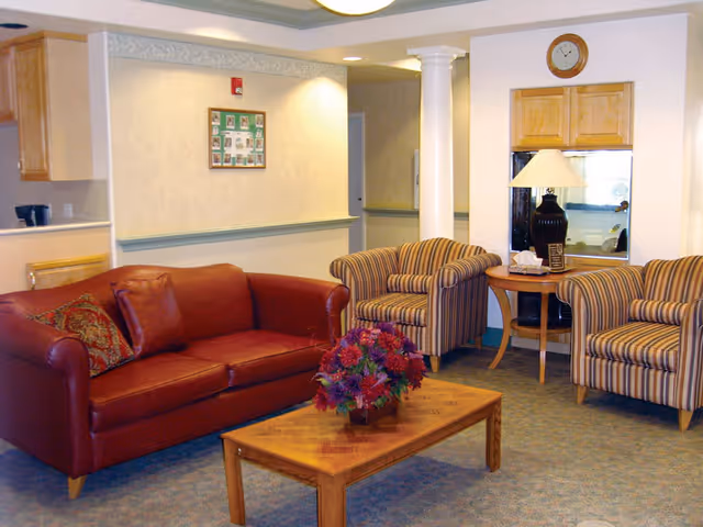 A cozy living room area with a red leather sofa, two striped armchairs, and a wooden coffee table with a flower arrangement. There is a side table with a lamp and tissues, a clock on the wall, and a framed photo collage. The room has beige walls and carpeted flooring.