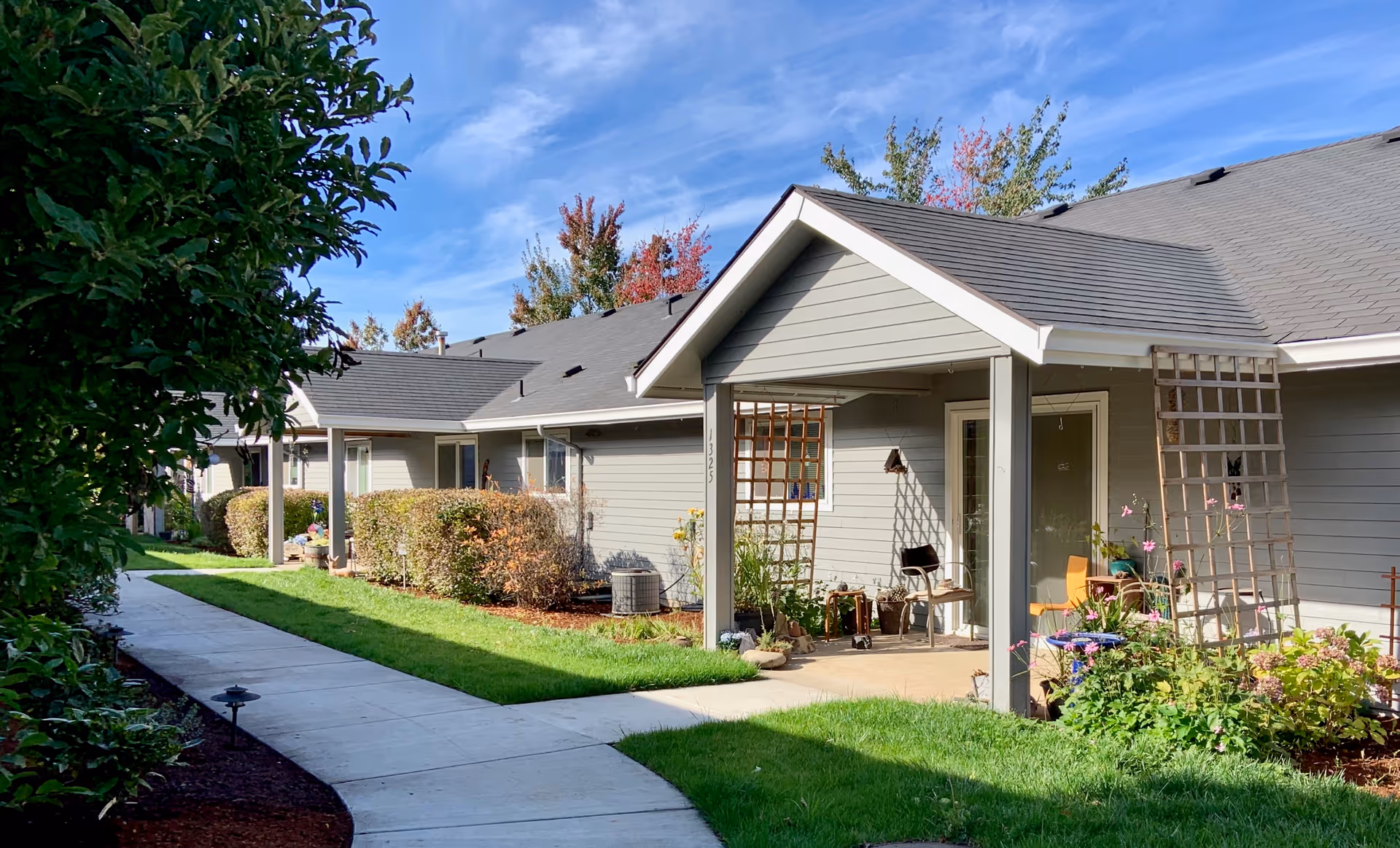Exterior view of a senior living facility showing a row of single-story units with gray siding and covered patios. The area features a concrete walkway, green grass, bushes, and some outdoor furniture and plants on the patios under a blue sky with light clouds.