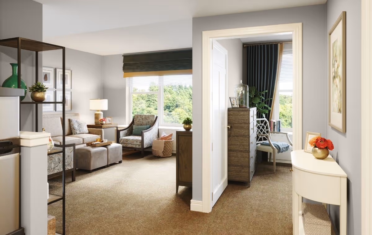 A cozy living area in a senior living facility featuring a beige sofa, two armchairs, a side table with a lamp, and two ottomans. The room has a large window with a green valance overlooking greenery outside. Adjacent to the living area is a small hallway leading to another room with a wooden dresser, a chair, and a window with dark curtains. The walls are painted light gray, and the carpet is beige. Decorative items include framed pictures, plants, and a vase with red flowers on a small white console table.
