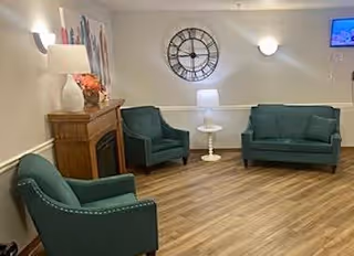 A cozy living room area with three teal upholstered chairs and a matching loveseat arranged around a small white side table. There is a wooden fireplace mantel with a lamp and decorative items on top. A large wall clock and a small wall-mounted TV are visible on the light-colored walls. The floor is wood with a light finish.