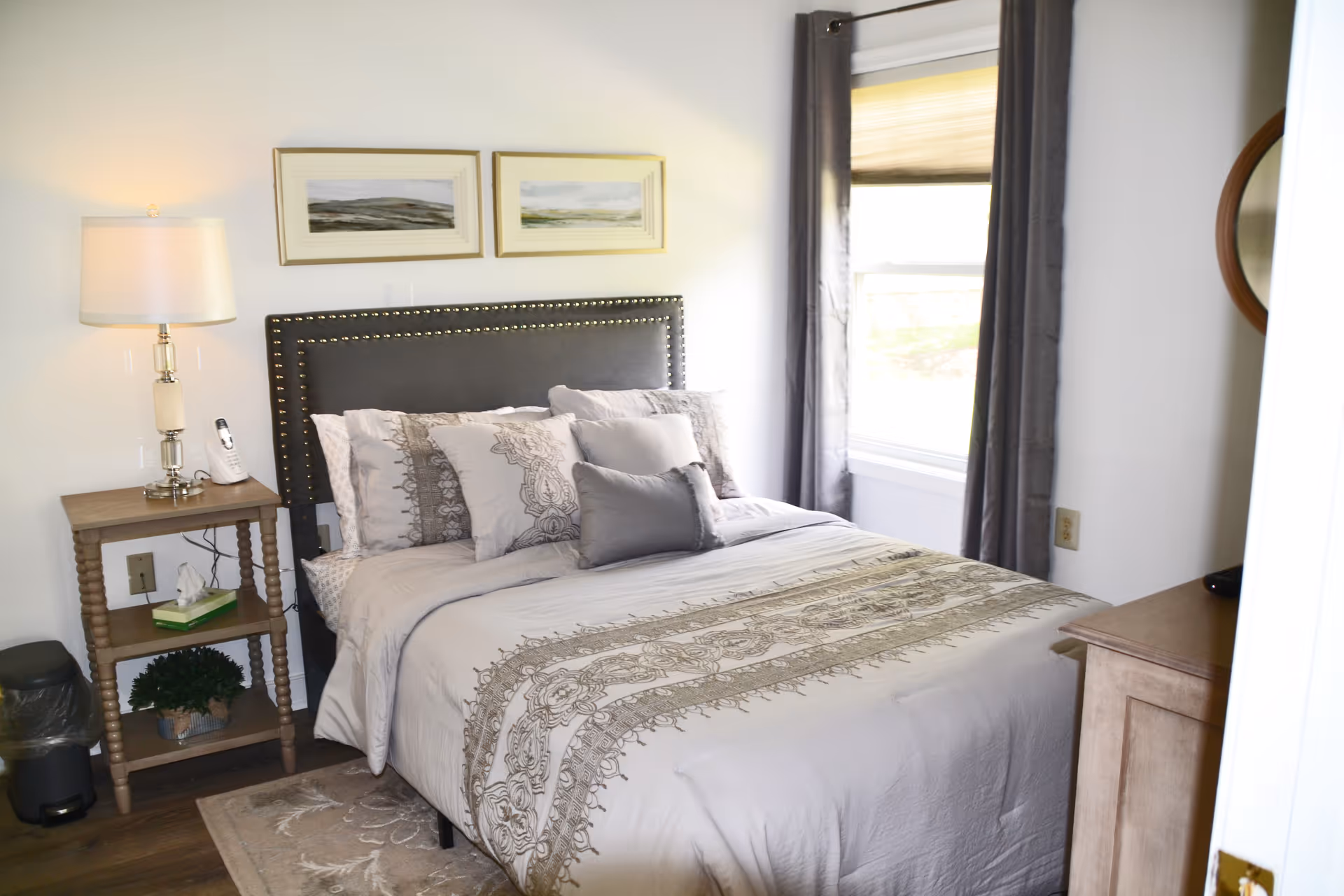 A neatly made bed with decorative pillows and a patterned beige comforter in a bedroom. Next to the bed is a wooden nightstand with a lamp, a cordless phone, a box of tissues, and a small potted plant. A window with gray curtains is on the right side, and a round mirror is partially visible on the wall.