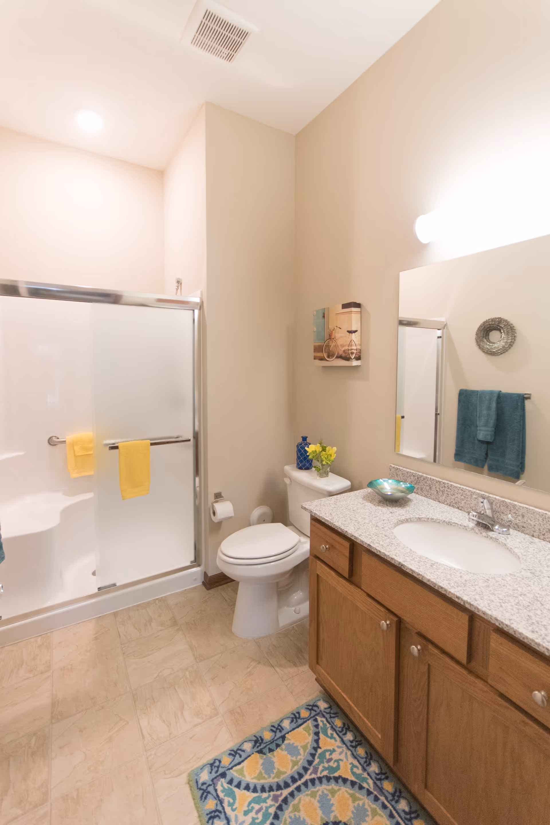 A clean and well-lit bathroom featuring a shower with sliding glass doors and yellow towels hanging on the handle, a white toilet with a blue vase and yellow flowers on the tank, a granite countertop with a sink, wooden cabinets below, a large mirror above the sink, a teal towel hanging on a rack, a decorative wall piece, and a colorful patterned rug on the tiled floor.