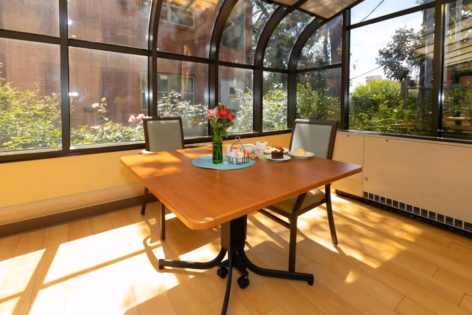 A sunlit dining area with a wooden table and two chairs. The table has a green vase with red flowers, a plate with a dessert, a plate with a bread roll, and some condiments. The room has large curved windows showing greenery outside.