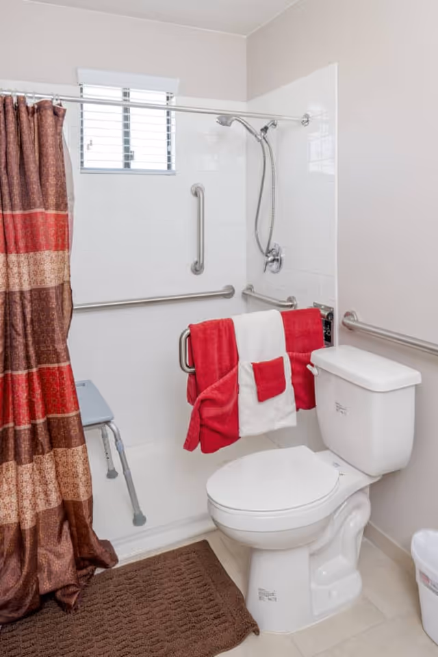Bathroom showing a toilet and a shower area with grab bars, a folding shower chair, patterned shower curtain, and red-and-white towels hanging on a grab bar.