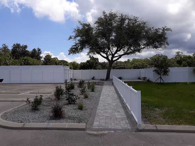 A paved walkway bordered by a white fence on the right and a landscaped area with small plants and gravel on the left, leading to a large tree in front of a white privacy fence under a partly cloudy sky.
