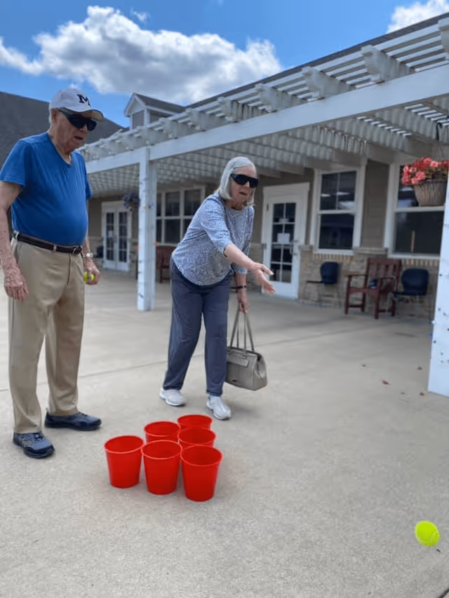Two elderly individuals playing an outdoor game with large red plastic cups arranged on the ground. One person is in the act of tossing a tennis ball towards the cups while the other watches. They are standing on a concrete patio area in front of a building with white pergola and windows.