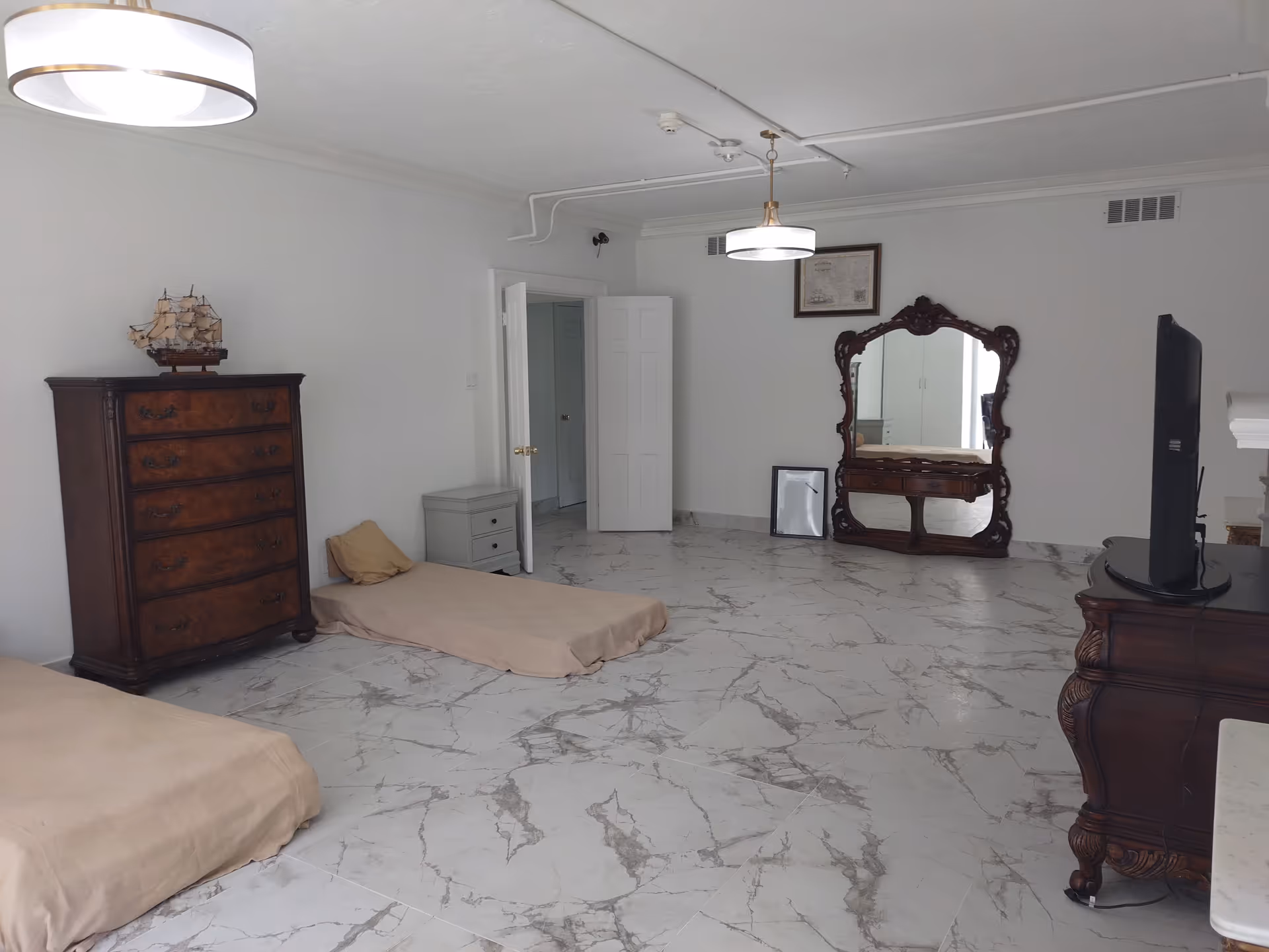 A spacious room with marble-patterned tile flooring, two low beds with beige covers, a wooden dresser with a model ship on top, a small gray nightstand, an ornate wooden framed mirror leaning against the wall, a flat-screen TV on a wooden stand, and two ceiling lights.