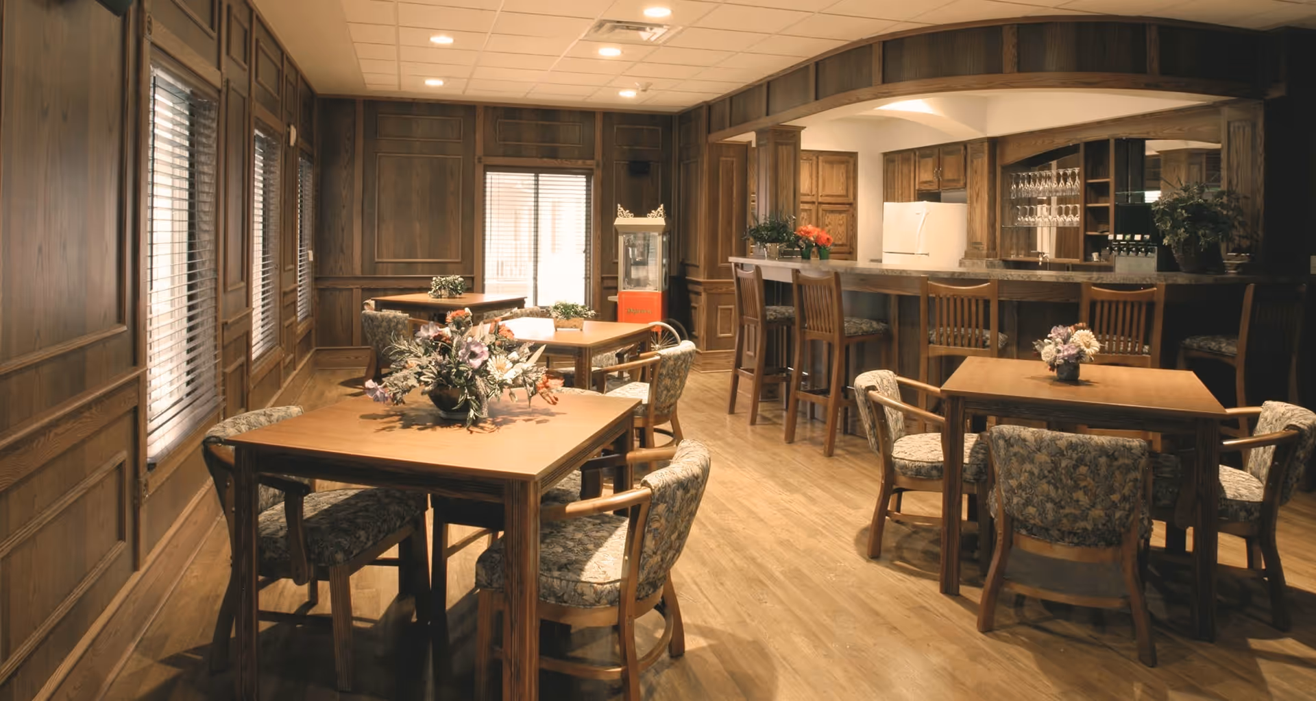 A cozy dining area with wooden tables and floral upholstered chairs. The room features wood-paneled walls and large windows with blinds. There is a bar counter with high chairs, a refrigerator, and glassware displayed behind the counter. A popcorn machine is visible in the corner, and floral arrangements decorate the tables.