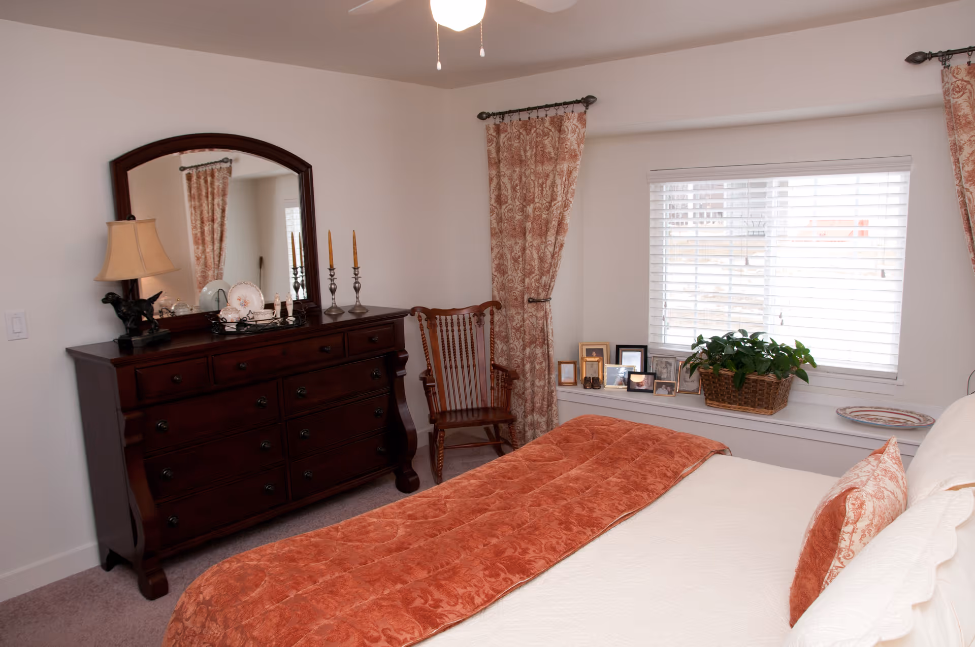 Light-filled bedroom with a bed topped by a rust-colored throw, a dark wooden dresser with mirror, a rocking chair, and a window seat with framed photos and a potted plant.