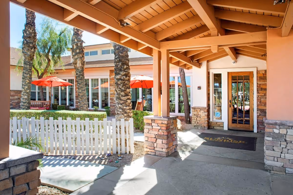 Covered entrance area of a senior living facility with stone pillars and wooden beams. There is a glass door with a wooden frame leading inside. Outside, there are palm trees, a white picket fence, green bushes, and red patio umbrellas providing shade to outdoor seating areas.