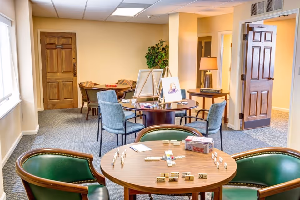 A cozy senior living common area with several round tables and chairs. One table in the foreground has a domino game set up with tiles and a box. In the background, there are more tables with chairs, some with art easels and paintings. The room has beige walls, carpeted floors, wooden doors, and a table lamp on a side table.