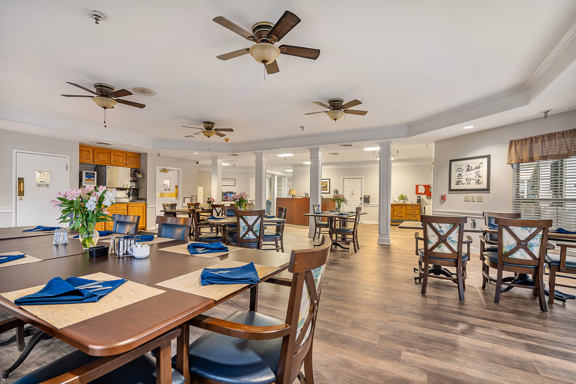 A spacious dining room with multiple wooden tables and chairs arranged neatly. Each table is set with blue napkins, placemats, and silverware. There are flower vases with pink and white flowers on the tables. The room has ceiling fans, wooden flooring, and large windows with blinds allowing natural light to enter. The walls are light-colored with framed artwork and a small kitchen area visible in the background.