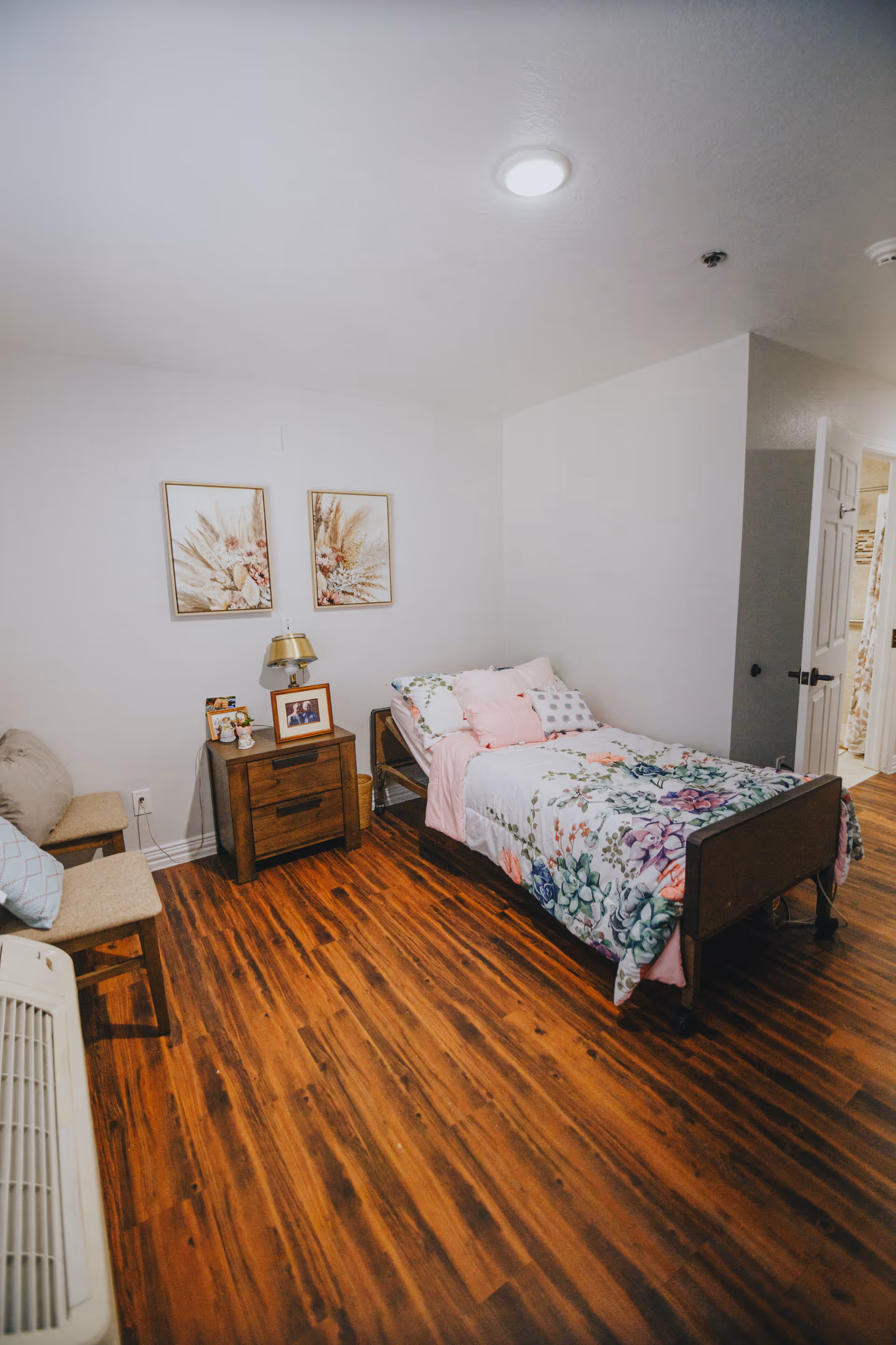 A cozy bedroom with a single bed featuring floral bedding and multiple pillows. Next to the bed is a wooden nightstand with a lamp, framed photos, and small decorative items. Two framed botanical prints hang on the white wall above the nightstand. The room has wooden flooring, a chair with a cushion, and an open door leading to another room.