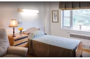 A single bedroom in a senior living facility with a bed covered in a striped bedspread, a wooden nightstand with a lamp and a small vase of yellow flowers, a comfortable armchair, a framed picture on the wall, and a window with a view of greenery outside.