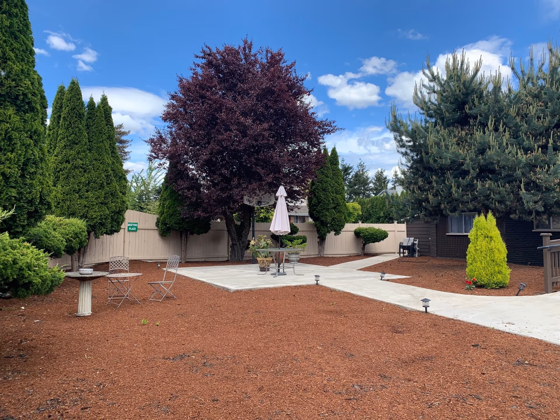 Outdoor garden area with a concrete patio featuring a table with an umbrella and chairs. The space is surrounded by various trees and shrubs, with a clear blue sky and scattered clouds above. A sign on the fence reads 'Meeting Place'.