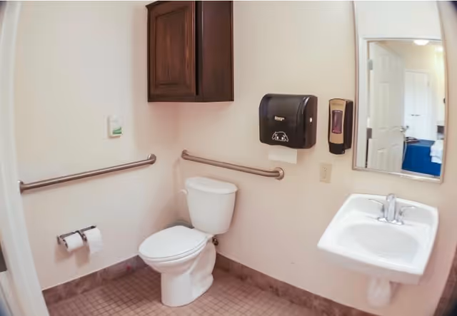 A clean and accessible bathroom with a white toilet, a white sink with a silver faucet, a wall-mounted paper towel dispenser, and a soap dispenser. There are grab bars on the walls near the toilet and sink. A wooden cabinet is mounted on the wall above the toilet, and a mirror is positioned above the sink. The floor has small square tiles in a neutral color.