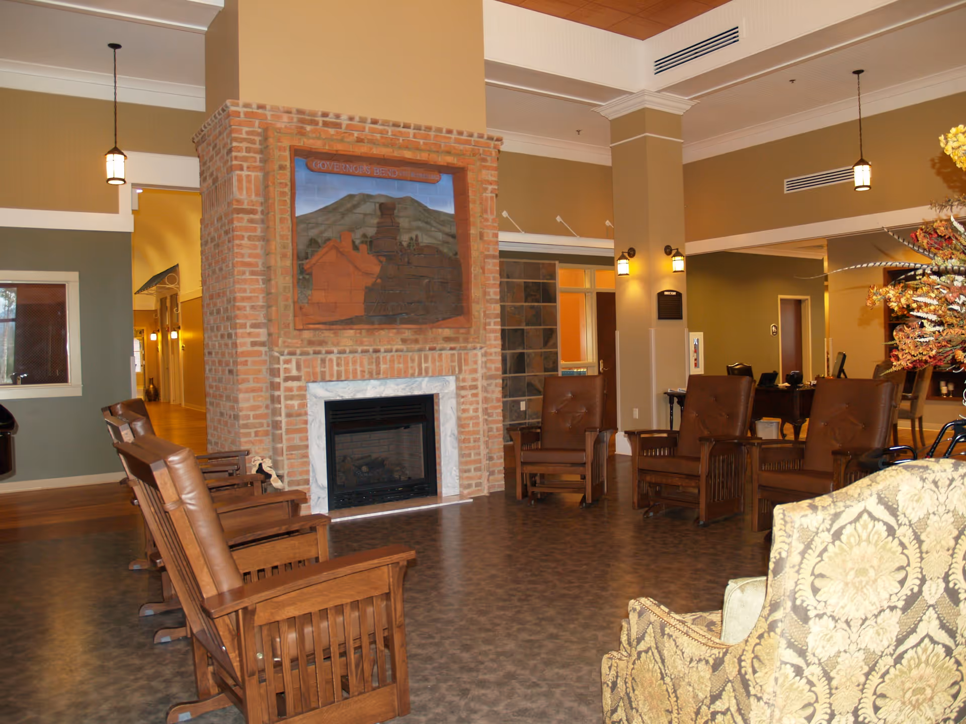 Interior view of a retirement and assisted living facility lounge area featuring a brick fireplace with a decorative tile mural above it. The room has several wooden chairs with brown cushions arranged around the fireplace, pendant lights hanging from the ceiling, and a patterned armchair in the foreground. The walls are painted in warm tones with white trim and there is a hallway visible in the background.