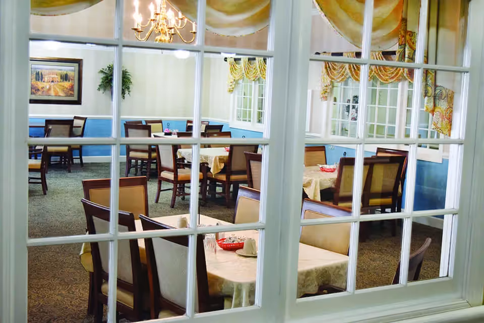 View through white-framed glass doors into a dining room with multiple tables covered in beige tablecloths and surrounded by wooden chairs with beige cushions. The room has blue wainscoting, beige walls, patterned carpet, a chandelier, framed artwork, and windows with yellow and patterned valances.