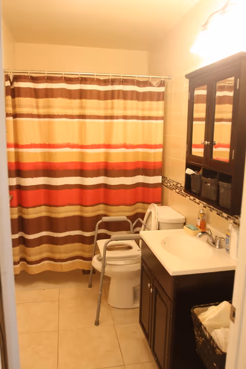 Small bathroom with a striped shower curtain, a toilet fitted with a raised seat and safety frame, a sink vanity and mirrored cabinet.