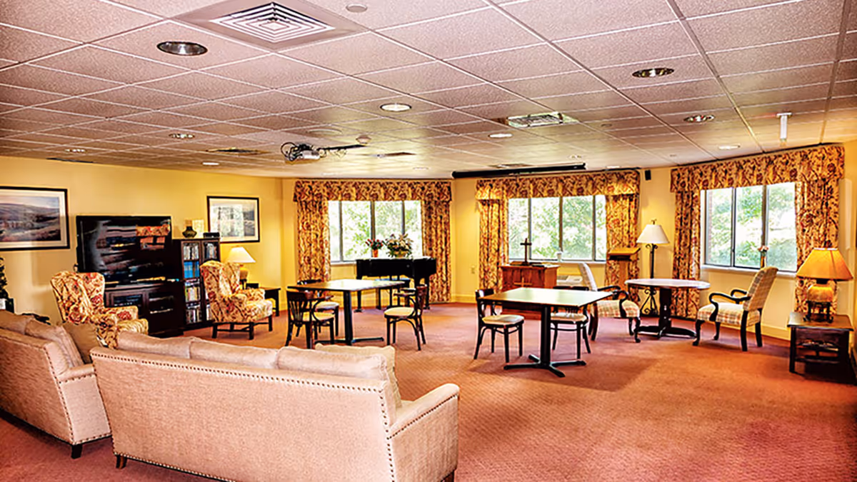 A spacious living room area with beige sofas, patterned armchairs, several tables with chairs, large windows with floral curtains, lamps, framed pictures on the walls, and a piano in the background.