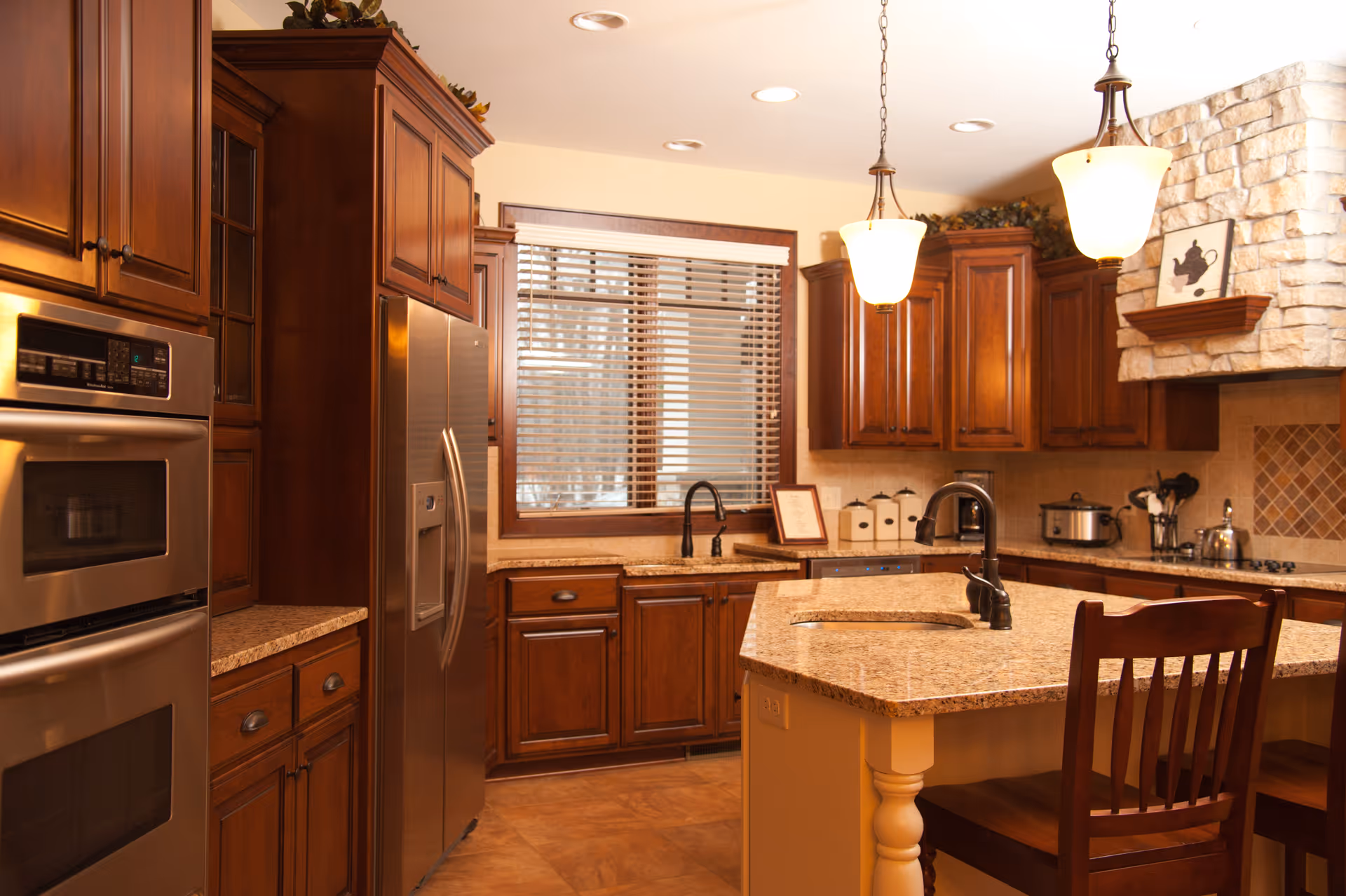 A warm, well-lit kitchen featuring wooden cabinets, a stainless steel double oven and refrigerator, granite countertops, a kitchen island with a sink and two wooden chairs, pendant lights hanging from the ceiling, and a window with blinds.